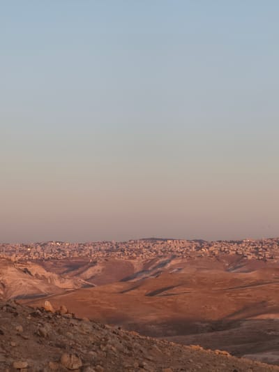 The image depicts a vast, arid landscape under a pale blue sky that transitions to a warm pink hue near the horizon. The foreground is dominated by rocky, barren terrain with scattered boulders and pebbles. In the mid-ground, rolling hills and valleys, characterized by dry earth in shades of brown and ochre, are visible. A distant city, with densely packed buildings and illuminated windows suggesting evening or dusk, sprawls across the horizon. The city appears to be built on a hilly terrain, with structures appearing as small, light-colored dots against the darker landscape. The overall impression is one of a remote, undeveloped area with a burgeoning urban settlement in the distance.