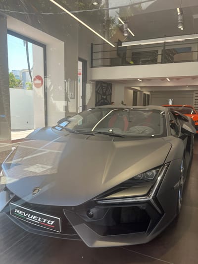 This media file captures a close-up, high-angle shot of a sleek, dark grey Lamborghini Revuelto parked indoors, likely in a showroom or dealership. The car's aggressive design is prominent, with its sharp lines and angular headlights clearly visible. The interior of the car, with its red accents, is partially visible through the windshield.
Reflections on the car's surface reveal elements of the surrounding environment. These include interior lighting fixtures, what appear to be glass doors or windows, and hints of foliage and outdoor structures, suggesting the car is displayed near a large window or in a space with outdoor visibility. One reflection shows a red and white circular "stop" sign, and another shows a palm tree, reinforcing a Mediterranean setting.
Text visible on the car includes the "REVELTO" model name, displayed prominently with the Italian flag colors on a rectangular badge. A Lamborghini crest is also visible on the car's bodywork. Other visible text is on signage in the background, appearing to be in Spanish, and includes "PLAZAS".
The lighting suggests it is daytime, with natural light filtering in from outside, creating a bright and reflective environment. The overall scene conveys a sense of luxury and automotive performance, characteristic of a high-end car dealership. There are no people clearly visible in the frame.