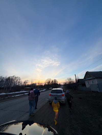 The image depicts a street scene in Дулесово, Russia, at dusk or dawn. The sky is a gradient of blue to orange near the horizon. A two-story building is visible in the background. A white car is parked on the side of the road. Several people are standing near the vehicle. One individual wears a yellow jacket. Another appears to be examining the car's rear. The road has cracks and minor damage. Bare trees line the horizon. Another vehicle's hood is visible in the bottom right corner.