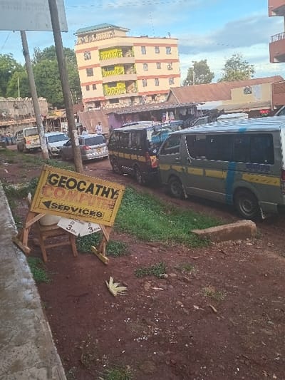 A yellow sign that reads "GEOCATHY COMPUTER SERVICES" sits on a wooden easel in front of a building in Karuri, Kenya at coordinates -0.72 latitude and 37.16 longitude. The sign is facing the street, which is unpaved and has a dirt and gravel surface. The sign is surrounded by green grass, with a small wooden stool to the left and a piece of palm frond on the ground to the right. There is a light brown building across the street with yellow window frames. The building is visible from the street. Behind the sign, there are several vehicles parked along the side of the street, including two white vans, one blue van, and several cars. Some people are walking around the street and the parking lot.  A few trees are visible in the distance.  The sky is a light blue with some clouds.