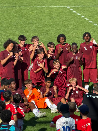 Here is a detailed description of the image:
**General Description:**
The image is an aerial shot of a group of people lying down on a grassy field, likely a soccer field, possibly after a game or training session. The image appears to be taken in bright, sunny conditions.
**People:**
The group consists of about fifteen to twenty people, mostly young males, likely teenagers. Most of them are wearing maroon athletic shirts with a logo that says "SS Etienne" and "MIFA", indicating they are part of a soccer team. The team appears to be diverse in terms of ethnicity. Some are wearing white shorts and socks. One person is wearing an orange shirt. There are others wearing red shirts and some in striped white and blue.
**Location:**
The image appears to be outdoors on a well-maintained soccer field. The city is Fentange, Luxembourg.
**Objects:**
-Soccer field with white lines marking boundaries.
-Athletic clothing (maroon shirts, white shorts and socks).
-Some individuals wear hats.
-A soccer jersey with "MBAPPE 10" written on the back.
**Actions/Interactions:**
The group is lying down on the grass, close together. Some are looking up at the camera and making hand gestures, like peace signs or thumbs up. They appear to be celebrating.
**Environment:**
The environment is a grassy field, likely a soccer field, with distinct white lines. The image suggests a bright, sunny day.