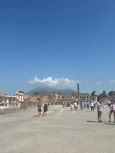 In the foreground of the image, a clear blue sky dominates the upper portion. Below, a vast, open, dusty plaza is populated by a diverse group of tourists. Many are dressed in casual summer attire, suggesting a warm climate. In the mid-ground, the ancient ruins of Pompeii are visible, characterized by weathered stone structures, columns, and arched doorways. Among these ruins, more people can be seen exploring. In the background, the imposing silhouette of Mount Vesuvius looms, partially obscured by a fluffy white cloud. The overall scene captures a moment of historical exploration on a bright, sunny day.