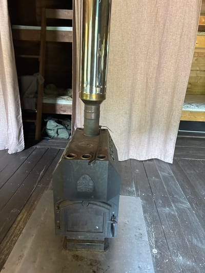 The image depicts the interior of a rustic room with wooden floors and walls, likely a cabin or a simple dwelling. In the center of the frame is a dark metal wood-burning stove with a shiny metal chimney pipe extending upwards. The stove has a rectangular firebox door with a handle. To the left of the stove, a pale pink curtain hangs, partially obscuring a set of wooden bunk beds. A green backpack is visible on the floor beneath the lower bunk. To the right of the stove, another pale pink curtain is visible, and beyond it, hints of a wooden wall and possibly another bunk bed can be seen. The overall impression is one of a simple, functional living space.