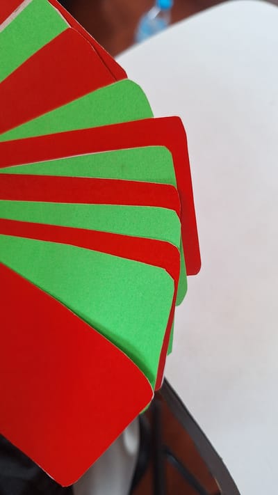The image shows a fan of alternating red and green cards or paper. The cards are fanned out in a curved pattern, with the edges overlapping. The surface they are on appears to be a white table. In the background, a blurry blue object, possibly a bottle of water, is visible. The image is taken from a close-up, angled perspective, focusing on the cards. The scene is indoors and the lighting suggests it is daytime. There are no people or animals visible in the image. It is not possible to determine the location in Karawang, Indonesia based on the visual information provided.