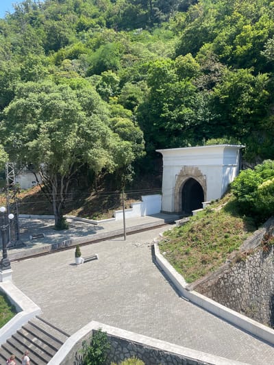 In Abaata, Abkhazia, a large, arch-shaped tunnel entrance, framed by light-colored stone and topped with a white facade, dominates the right side of the image. The tunnel is dark inside. The entrance is situated on a grassy, sloped embankment covered in vegetation. To the left of the tunnel, a paved area transitions to a railway track. A lone tree with a thick trunk and expansive branches stands in the middle ground, casting shadows. Further left, a black lamppost with two lights and a metal structure are visible. The ground is paved with gray interlocking bricks. In the foreground, a concrete staircase leads down, with two individuals partially visible at the bottom. The scene is set against a backdrop of a dense, verdant forest covering a steep hillside under a clear blue sky.