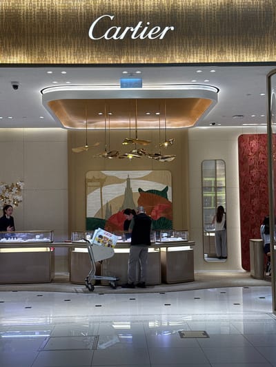 In a brightly lit, modern interior, possibly a luxury boutique, a person is seen pushing a metal shopping cart. The cart has a white bag with colorful cartoon characters and text that appears to say "POP MART". The person pushing the cart is wearing dark clothing. In the background, the store displays are visible, with illuminated shelves and glass cases. The store's signage is prominently displayed on the left side of the image, featuring the word "Cartier" in white, stylized lettering against a textured, gold-toned background. The overall ambiance suggests a retail environment, likely during daylight hours given the interior lighting. The scene appears calm and orderly. The location is identified as Nong Prue Subdistrict, Thailand.