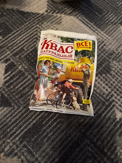 The image shows a close-up, slightly angled shot of a white and yellow package of kvass mix, set against a dark gray, textured surface that appears to be a blanket. The package features vintage-style illustrations.

The front of the package displays the word "КВАС" (KVAS) in large red letters. Below this, in smaller red letters, is "НАТУРАЛЬНЫЙ" (NATURAL). A yellow banner runs across the top, containing the text "ВСТ" (ALL) and "ВКЛЮЧЕНО" (INCLUDED) in black text. Beneath the main title, there is a line of text in a smaller font that reads "ГОТОВАЯ СУХАЯ СМЕСЬ. ДОБАВИТЬ ТОЛЬКО ВОДУ" (READY DRY MIX. JUST ADD WATER).

The central illustration depicts several figures. On the left, a woman in a light blue dress is holding a glass to her lips, seemingly drinking. Next to her, a person in a white hat and uniform is attending to a large yellow tank truck, holding a jar. In front of the truck, a young boy on a bicycle is reaching towards a golden-brown dog that is sitting on the ground. A man in a military-style uniform and cap is standing near the truck, looking towards the boy and the dog, and holding a bottle. The truck has the word "Кіѕ" (KIS) written on its side.

At the bottom of the package, a yellow strip contains the text "НА 2 Л" (FOR 2 L) and further to the left, text that appears to read "«РЕЦЕПТЫ ВКУСА» - ВКУСНО, КАК РАНЬШЕ!" (“RECIPES OF TASTE” - DELICIOUS, LIKE BEFORE!).

The image does not contain any visual cues that would allow for its specific location in the city of Chernogorsk, Russia, or any other specific urban environment. The background is a plain, textured fabric.
