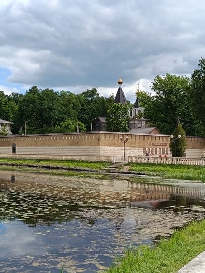 The image captures the Pavlovskoye Gidrogeologicheskoye Lyceum (Koltushi, Russia) on a cloudy day. In the foreground, a body of water is visible, covered with lily pads and reflecting the surrounding environment. A stone wall with a lighter stone base borders the water, topped with a decorative railing. Behind the wall, a religious building with multiple domes, characteristic of Russian Orthodox architecture, stands amidst lush green trees. A memorial plaque is mounted on the wall. A lone figure can be seen walking on the path behind the railing. The sky is overcast with large, dark clouds interspersed with patches of blue sky.