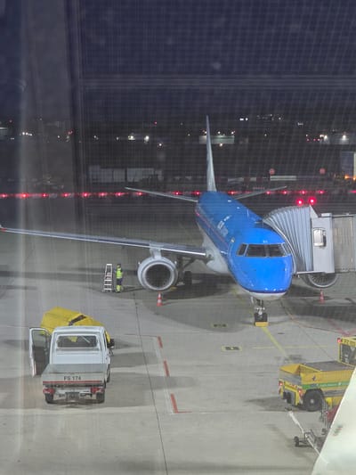 This is a vertical, slightly angled, nighttime aerial shot of an airport tarmac in Echterdingen, Germany, seen through a window. A bright blue KLM airplane is connected to a jet bridge. The airplane has "KLM" and "The Flying Dutchmen" written on its side. A small white truck with its driver's side door open is parked near the wing of the plane. The truck has "FS 174" written on its tailgate. A person in a yellow vest and green pants stands near a ladder next to the airplane's engine. A yellow covered object is also visible near the truck. In the background, there are multiple red lights on a fence, and lights from airport buildings and structures. Another yellow vehicle, a baggage tug, is visible on the right side of the image. The tarmac is marked with yellow and red lines.