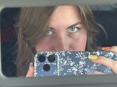 The image shows a close-up reflection in a mirror of a young Caucasian woman with light brown hair and blue eyes, looking directly at the camera. She has freckles across her nose and cheeks. She is holding a smartphone with a floral case in front of her face, obscuring her mouth and chin. Her nails are painted with colorful designs, including yellow, blue, and orange. The background is dark and out of focus, suggesting an indoor setting, possibly a car interior. The image does not contain any specific elements that would indicate its location in the city of Можга, Russia.