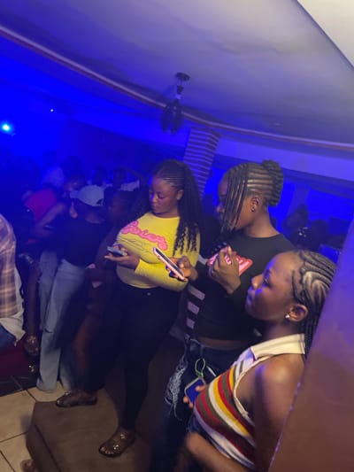 A group of young Black individuals are gathered in a dimly lit, indoor space, likely a club or lounge, in Agwala, Nigeria. Blue lighting illuminates the scene, creating a vibrant and energetic atmosphere. In the foreground, three young women are the primary focus. The woman on the left, wearing a yellow long-sleeved shirt with the word "POSH" in pink lettering, is holding a smartphone and appears to be looking at it. To her right, a woman in a black long-sleeved shirt with a red and pink graphic on the chest is also looking downwards, her hands clasped together. The third woman, on the far right, is partially visible, wearing a striped tank top and looking to her right. The background shows more people in silhouette, some appearing to be dancing or socializing. The environment suggests a nightlife setting, with subtle hints of brickwork on a wall behind the central figures and a tiled floor.
