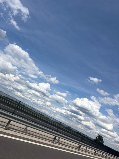 The image shows a view from a bridge or overpass, likely in Óföldeák, Hungary, based on the request. The sky is predominantly blue with scattered cumulus clouds. A section of roadway with a metal guardrail is visible in the foreground. Beyond the roadway, there is a view of what appears to be farmland.