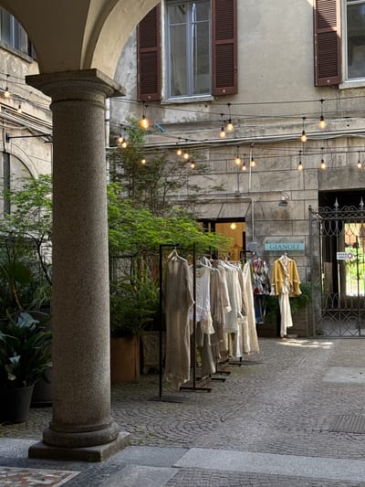 The image depicts an outdoor clothing display in a narrow alleyway, characteristic of the city of Como, Italy. The display, positioned near the "GIANOLI" store, consists of multiple racks filled with light-colored clothing items. A stone pillar, part of an arched structure, dominates the foreground, obscuring a portion of the view. String lights hang overhead, creating a warm ambiance. The alleyway is paved with cobblestones, adding to the historical atmosphere. Vegetation, including trees and potted plants, is interspersed throughout the scene, softening the urban environment. No people are visible in the immediate view.