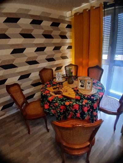 The image depicts an interior domestic space, likely a dining area, located in Wolfisheim, France. The floor is covered with light-toned laminate flooring arranged in planks. A wall on the left features geometric wallpaper with a repeating three-dimensional block pattern in shades of black, light grey, and beige.
A round table, draped with a dark tablecloth adorned with red and orange floral and fruit patterns (resembling pomegranates and blossoms), occupies the center of the frame. Centered on the table, a dark vase holds several pink flowers. To the left of the vase, a white decorative clock with a heart motif is visible. To the right of the vase, a folded wooden fan with dark and gold designs rests on a placemat. Further right, a wooden double photo frame containing two indistinct images is present, also on a placemat.
Five wooden chairs with woven cane seats and backrests are arranged around the table. One chair in the immediate foreground is seen from behind, facing away from the table. A large window or glass door is present on the right side, partially covered by a single panel of a plain orange curtain. Exterior roller shutters are visible through the window panes, appearing partially lowered. No individuals are present in the scene, and no actions or interactions are depicted.