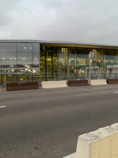 The image shows the exterior of a modern building with a glass facade, likely an airport terminal, in Manises, Spain. The building has multiple floors with reflective windows that show internal lights and structures. A metal fence runs along the front of the building, separating it from a row of concrete barriers and a paved road. The sky is overcast, suggesting a cloudy or twilight condition. There are no visible people or vehicles in the scene. The concrete barriers appear to be placed to delineate traffic areas or for security purposes.