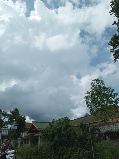The scene is set in Cibadak, Indonesia. The upper two-thirds of the image features a sky with a mix of white and grey cumulus clouds, with some clear blue sky visible. In the lower-left quadrant, three individuals are seated on a white scooter. The driver, an adult female wearing glasses and a patterned brown hijab, operates the vehicle. Behind her, another adult female with glasses is seated. A child in a light-colored shirt is positioned in front of the driver, looking towards the viewer. The scooter's license plate is partially visible, starting with the letter 'F'. The middle ground contains multiple buildings with green-pitched roofs and light-colored walls, largely obscured by dense green foliage and trees. A metal fence runs in front of these structures. A vertical white sign, partially visible on the far left behind foliage, displays text including "MELATI 1", "KLAS J", and "MAS". The overall environment indicates a daytime, semi-urban or suburban setting.