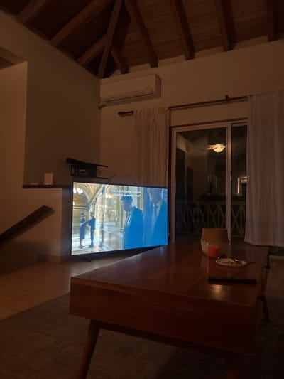The scene depicts a dimly lit interior living area within Olde Yard Village, British Virgin Islands, observed under low ambient light conditions. A large, flat-screen television is centrally positioned on a low stand against a light-colored wall, displaying a moving image. The screen shows three figures, two males in dark suits and one female in a dark dress, within an architectural setting featuring arches. White subtitles are visible across the top of the screen, stating: "I can't do it, Dottila. Could we not pick it up another time?"
Above and to the left of the television, a dark, elongated object, possibly a soundbar, is mounted on the wall, beneath a small white wall shelf holding a white electronic device. In the foreground, a rectangular dark wooden coffee table is present, upon which rests a woven basket, a lit orange candle in a clear glass holder, a small white dish containing a metallic spoon and several small dark fragments, and a black remote control.
To the right of the television, a glass sliding door or window unit is visible, partially flanked by white curtains suspended from a wooden rod. The glass reflects an interior or exterior space featuring a ceiling-mounted light fixture and a balustrade. The room's architecture includes a high vaulted ceiling with exposed dark wooden beams and light-colored tiled flooring. A white wall-mounted split-unit air conditioning unit is affixed high on a wall. A section of a wooden staircase or railing is partially visible along the left wall.