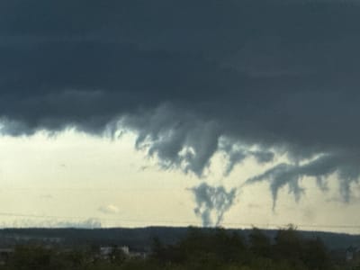 The image depicts a cloudy sky above the city of Lipaki, Russia. The foreground consists of a dark, indistinct mass of trees and possibly rooftops. Above this, a band of light grey separates the ground from the heavy, dark clouds looming above. These clouds display a ragged underbelly with wisps and tendrils of grey reaching downwards, suggesting an approaching storm. The dominant color palette is grey and dark blue.