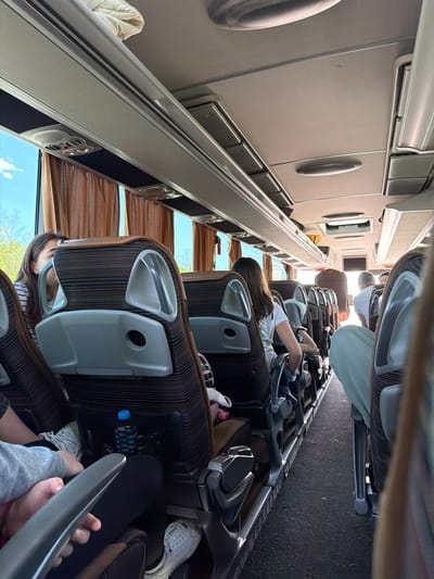 The image depicts the interior of a bus with passengers seated. Passengers are in the foreground and middle ground. The interior is dimly lit, with overhead compartments running along the left side of the frame. Brown curtains cover the windows. Seats are brown with a striped pattern and grey headrests. Passengers are in varying states of relaxation, with some looking out the windows. A water bottle is visible next to one of the seats. The top portion of the image shows luggage racks. The location in the city of Yazdach, Bulgaria, is not discernible from the image.
