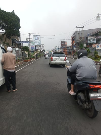 A street scene in the city of Chagzam_Pam, Bhutan, captured on an overcast day, depicts vehicles and pedestrians on a paved road. On the far left, a male individual wearing a white skullcap, light brown top, dark pants, and sandals stands beside a white vehicle, facing away from the camera. In the right foreground, a dark scooter with Indonesian license plate 'F 2830 WA' carries a helmeted driver and a passenger in a gray top and black leggings, wearing pink shoes. Further down the road, traffic includes a silver Toyota Innova displaying Indonesian license plate 'D 1709 ZA', among other cars. The left roadside features a stone wall with a sign reading 'BIARA St CLARA'. Utility poles with electrical wiring and various commercial billboards, including 'CLAS', 'ELITE', and 'STUDIO', line both sides of the street, which is flanked by multi-story commercial buildings extending into the background.