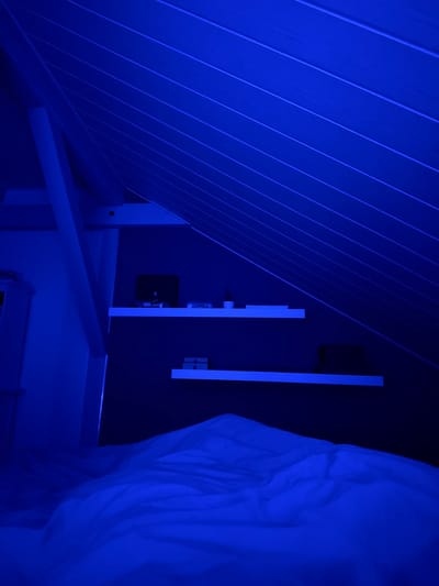 An interior scene is bathed in blue light. A wall with angled planking is on the left. Two vertical white panels are mounted on a darker wall, with what appears to be electrical outlets and a black rectangular device between them. A portion of a white, textured curtain or bedding is visible on the right, obscuring the background. Below, a white structure with a crossbeam and horizontal platform is visible.