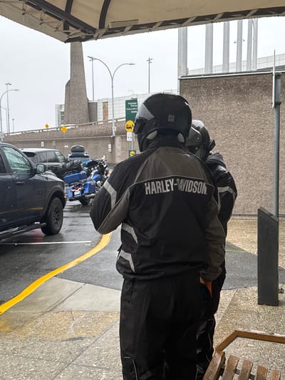 The image depicts two individuals wearing black motorcycle jackets with "HARLEY-DAVIDSON" prominently displayed in white lettering. They are on a paved surface, possibly a street or parking lot, with several motorcycles parked nearby, including a bright blue one. The scene appears to be outdoors, with a building structure visible in the background. A dark-colored pickup truck is parked in the foreground. The ground is wet, suggesting it might be raining or has recently rained. Yellow lines are painted on the pavement.

The image is oriented vertically, with the subjects appearing upside down relative to standard orientation. This suggests the photo was taken with the camera held in a non-standard position or that the image has been rotated. The sky is overcast, indicating a cloudy or rainy day.

The overall atmosphere seems to be one of preparation or a pause during a motorcycle ride, possibly in anticipation of inclement weather. There are no explicit emotional cues conveyed by the subjects.

Visible text includes "HARLEY-DAVIDSON" on the jackets. There are also some signs in the background, though the text on them is too small to read clearly. One sign appears to be for "Passenger Pick-up / Embarcadère".