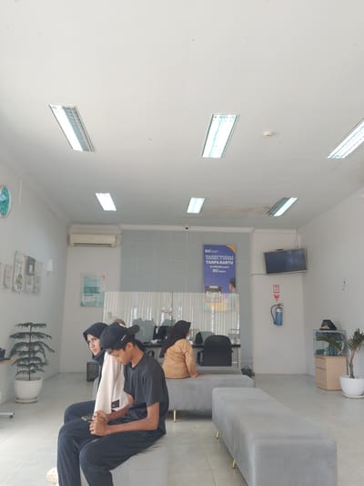 This image captures the interior of what appears to be a waiting area, likely a bank or service center, in Manetunong, Indonesia. Three individuals are visible, seated on grey upholstered benches. In the foreground, a young man wearing a black t-shirt and cap is engrossed in his phone, seated next to a young woman in a white top and black hijab, who is looking towards the left. Further back, another person in a light brown top and hijab is seated on a bench, facing away from the camera, behind a glass partition.
The setting features white walls and a light-colored tiled floor. The ceiling is white with several long, recessed fluorescent lights providing illumination, along with a visible smoke detector. A wall-mounted air conditioner is on the left wall. Various items adorn the walls: a round wall clock on the far left, several posters and flyers, and a prominent blue poster on the back wall. This blue poster clearly displays text: "BSI", "TARIK TUNAI TANPA KARTU", "di ATM BSI pakai", and "BSI mobile", indicating this is a branch of Bank Syariah Indonesia.
Other objects include a wall-mounted television screen that is off, a blue fire extinguisher below it, and a small wooden cabinet with a potted plant on the right side of the room. A large potted plant is also present on the left side near the foreground subjects. The scene suggests a calm, routine atmosphere of people waiting for a service.