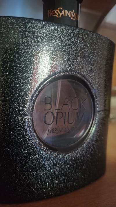 The image shows a close-up of a black, glittery perfume bottle, specifically Yves Saint Laurent's Black Opium. The bottle has a circular indentation on its front with the words "BLACK OPIUM" embossed in a darker shade. Below this, "YVES SAINT LAURENT" is also embossed. The bottle's surface is textured with numerous small, reflective specks, giving it a sparkling appearance. The top portion of the bottle, partially visible, displays the "YVES SAINT LAURENT" logo in gold lettering. The background is blurred but suggests a wooden surface and hints of a blue and brown environment. The location of this image in Dagomys, Russia, is not discernible from the visual information provided.