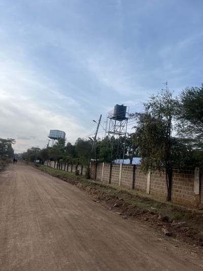 A long, unpaved road stretches into the distance under a cloudy sky. Tall trees and a block wall line the right side of the road. Water towers stand prominently on the left side, appearing twice in the frame. Power lines run parallel to the road. The scene evokes a sense of rural or semi-urban development.