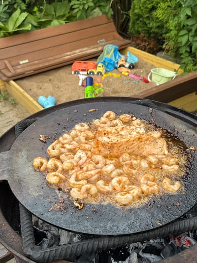The image shows a large, round, black skillet, likely a griddle or paella pan, filled with sizzling shrimp and some pieces of meat. The skillet is placed over glowing coals, indicating outdoor cooking, likely a barbecue. The shrimp are a light pinkish-orange color, some curled from the heat, and appear to be seasoned with spices. The meat, possibly chicken or fish, is a lighter brown color and is also cooking in the pan. The overall scene suggests a casual outdoor meal preparation.
In the background, to the left of the skillet, is a sandbox. Scattered within the sandbox are various children's toys, including a red toy truck, a blue toy dump truck, and a yellow toy vehicle with a trailer. There is also a light green bucket and a few other colorful plastic toys. The sandbox appears to be made of wood. Surrounding the sandbox and the cooking area are lush green leaves and foliage, suggesting a garden or backyard setting. The lighting indicates it is daytime, likely a clear day given the shadows.
The image was taken in Rezekne, Latvia. There is no visible text in the image. The focus is on the cooking activity and the surrounding environment.
