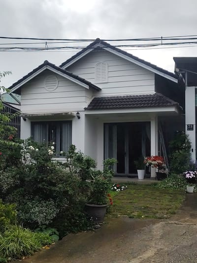 The image shows a two-story house with a white exterior and a dark tiled roof. The house is surrounded by lush greenery, including trees, bushes, and flowering plants. There are several potted plants on the porch and in the garden. A dark-colored pathway leads to the front door, which is partially obscured by plants. Overhead, power lines are visible against a cloudy sky. A house number, "73," is affixed to a pillar on the right side of the house. The scene appears to be in a residential area, possibly in the city of Lang Biang - Đà Lạt, Vietnam, known for its cool climate and floral landscapes.