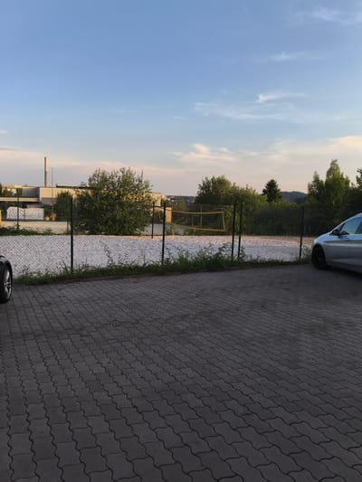 The image depicts a paved parking area in Pegnitz, Germany, with a volleyball court visible in the background. The sky is a clear blue with some wispy clouds. On the left side of the frame, a portion of a car's wheel is visible. In the mid-ground, a fenced-off area contains a sand volleyball court with yellow net poles. Behind the court, trees and industrial buildings are present. A silver car is parked on the right side of the paved area. The ground is covered in interlocking gray pavers.