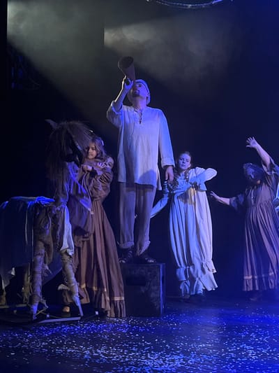 This image captures a theatrical performance on a stage in Novoshakhtinsk, Russia. Five individuals are present under blue-toned stage lighting, with a dark background and the stage floor covered in small white fragments resembling confetti or artificial snow.
Centrally, an adult male stands elevated on a dark, rectangular prop box, wearing a light-colored tunic, light trousers, and a cap. He holds a brown conical horn to his mouth, tilted upwards, as if making an announcement. To his immediate left stands a young female, approximately 6-10 years old, in a long, dark-brown dress with light trim, her hands clasped. Beside her, on the far left, is a rustic, light-colored donkey-shaped prop with a shaggy mane, mounted on a rocker base.
To the right of the central male, a young female, approximately 12-18 years old, stands in a light-cream long-sleeved dress with ruffled details, her right arm slightly raised and bent. Her gaze is directed forward. Further to the right and slightly behind, an adult female in a dark-brown long-sleeved dress performs a dramatic gesture with her left arm raised high and bent. A fifth individual, also in a dark-brown dress, is partially visible behind the gesturing female on the far right. The overhead lighting creates atmospheric haze and distinct shadows.