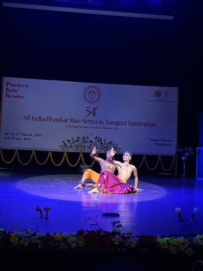 The image shows two men performing a traditional Indian dance on a stage lit by blue lights. The stage is decorated with a white banner that reads "All India Bhaskar Rao Nritya & Sangeet Sammelan" and "Unveiling the Soul of Indian Classical Arts"  in large black font. The banner also shows a picture of many performers. There is a red banner at the top with the words "Pracheen Kala Kendra" and "54th" in gold lettering. The stage has garland decorations along the edges, with the garland falling in arcs, a few feet above the floor. The stage is illuminated by purple lights.  The two performers are wearing brightly colored traditional costumes. The man on the left is wearing an orange dhoti and the man on the right is wearing a pink dhoti. They are both shirtless and have their arms raised in a dance pose. The background features the "Tagore Theatre Chandigarh" sign. The image appears to be taken from the audience, with the flowers adorning the front of the stage being visible in the foreground. The coordinate location 30.733, 76.789 corresponds to Tagore Theatre in Chandigarh.