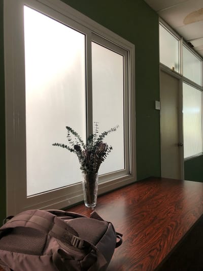 The image shows a room in Agia Paraskevi, Greece, located at 37.998 latitude and 23.82 longitude. The room is painted a dark green and features a large window with a white frame. The window is divided into two panes. The window provides a view of a cloudy sky. Outside the window, a white door can be seen closed. A vase of dried plant life sits on a wooden table in the foreground. A brown backpack is leaning against the table. The top of the table appears to be made of dark wood grain. The table is in the bottom right corner of the image. The backpack is in the bottom right corner of the image, partially out of frame. There are no people in the image.