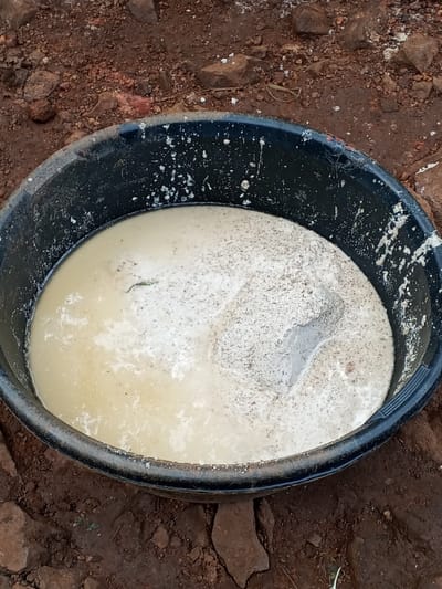 The scene is located in Getenga ward, Kenya. A black, circular plastic basin, approximately 60-80 cm in diameter, is centrally positioned on reddish-brown, dry earth. The basin contains a heterogeneous mixture. The left side of the basin is filled with a pale yellow-beige liquid. The right side contains a mound of light grey, granular material, heavily speckled with numerous dark brown to black particles, which appears partially saturated by the liquid. A small, dark green leaf fragment, approximately 3-4 cm in length, is partially submerged in the liquid on the left. White, dried particulate splatters are visible on the interior upper rim and exterior upper portion of the basin. The surrounding ground is unpaved, consisting of dry, reddish-brown soil intermingled with various sizes of dark brown, reddish, and grey rocks and pebbles. Scattered lighter, possibly white, particulate matter is present on the soil near the top edge of the frame. No individuals are visible.