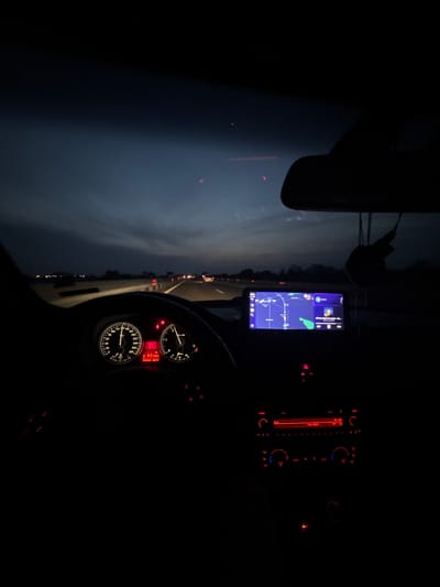 The image shows the interior of a car at night while driving on a highway near Opalchenets, Bulgaria. The dashboard is visible, with illuminated gauges showing the speedometer and RPM. A smartphone is mounted in the center console, displaying a navigation app with a map. The car radio and climate control panel are lit up in red. Outside the car, a highway is visible with other cars' tail lights. The sky is dark.