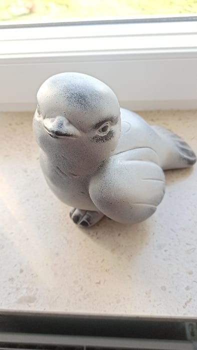 A light gray, textured bird figurine with darker speckling is positioned indoors on a light-colored, speckled windowsill in Florenville, Belgium. The figurine, approximately 15-20 cm in length, faces slightly left, displaying stylized eyes, beak, and wing contours. It casts a subtle shadow to its right. Directly behind the figurine is a white, multi-pane window frame. Beyond the window, a blurred outdoor environment with green foliage is visible under natural light. No individuals are present in the scene, and no specific actions or interactions are depicted.