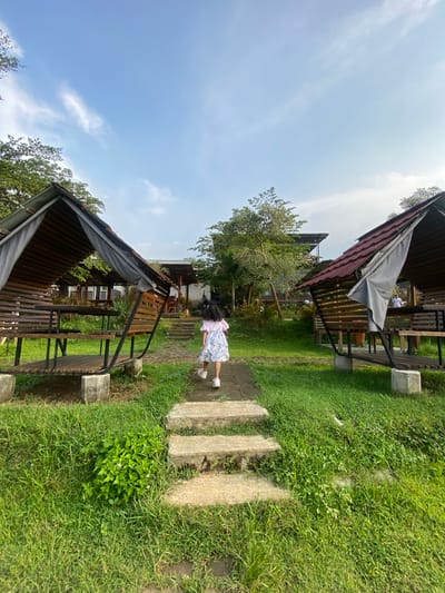 A young girl walks on a stone path between two wooden structures with roofs in Penanggungan, Indonesia. The structures are surrounded by grass and trees under a partly cloudy sky.