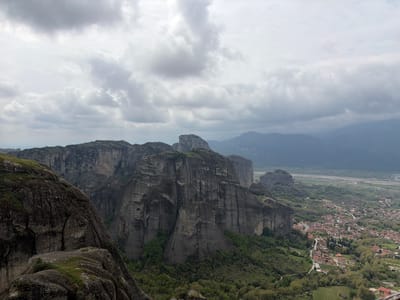 The image captures a high-angle, wide view of the landscape surrounding the town of Kastraki in Greece. Dominating the scene are the unique rock formations of Meteora, characterized by their towering, vertical cliffs and unusual shapes. The rock formations are a mix of gray and dark gray hues, with patches of green vegetation clinging to their bases and crevices.
Below the rocks, the town of Kastraki is visible, featuring clusters of buildings with red-tiled roofs, interspersed with trees and gardens. Roads wind through the town, connecting the buildings and leading into the surrounding valley. In the distance, a broad plain stretches out, with hints of a river and possibly other settlements. Mountains rise in the background, fading into a hazy blue-gray color, indicating distance.
The sky is overcast, filled with thick, gray clouds that diffuse the light, creating a somewhat muted and soft overall atmosphere. There is no visible human activity, and the overall impression is one of tranquility and natural grandeur.