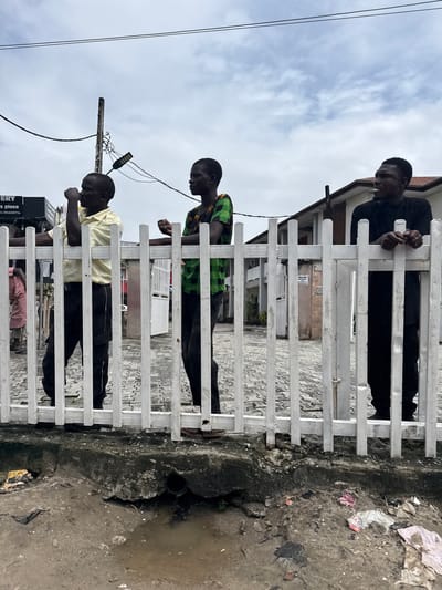 Three young men are standing behind a white picket fence, looking out. The man on the left is wearing a yellow short-sleeved shirt and black trousers, with his right arm resting on the fence and his left hand near his chin. The man in the middle is wearing a green and black checkered shirt and black trousers, with his arms crossed. The man on the right is wearing a black t-shirt and black trousers, with his hands clasped in front of him. The fence is positioned in front of a paved area and a building. In the foreground, there is a dirty, uneven ground with debris and a puddle of water, indicating a less maintained area. The sky above is overcast with clouds. The overall impression is a scene in an urban environment, possibly a street in Lagos, Nigeria, where the contrast between the fenced-off area and the surrounding environment is visible.