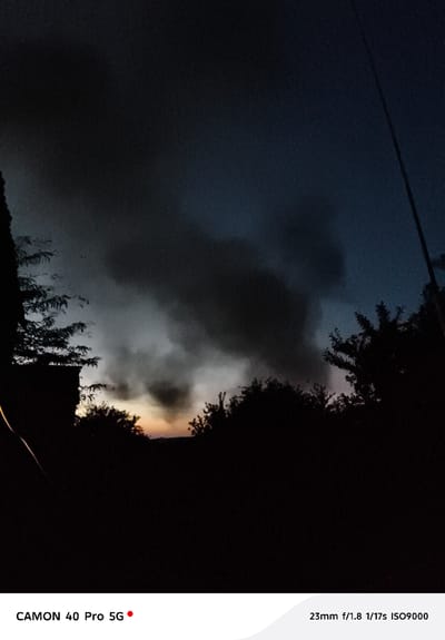 This is a vertical, low-light photograph taken at dusk in Kramatorsk, Ukraine, during what appears to be stormy weather. The foreground is dominated by a silhouette of trees and possibly buildings, creating a dark, indistinct landscape. The sky is a mix of deep blues and grays, with a large, dark mass of smoke or storm clouds occupying the upper portion of the frame. This dark formation is dramatically lit from below by a sliver of orange and yellow light on the horizon, indicating sunset. The overall mood is somber and dramatic, possibly conveying a sense of unease or the aftermath of an event.
Visible text at the bottom of the image indicates camera settings and model information: "CAMON 40 Pro 5G" and "23mm f/1.8 1/17s ISO9000". The camera settings suggest a very low light environment, with a relatively long exposure time and high ISO, which is consistent with the dark, grainy quality of the image. The presence of smoke or storm clouds, combined with the location context of Kramatorsk, Ukraine, suggests the possibility of a recent conflict or industrial incident, though this cannot be definitively determined from the image alone.