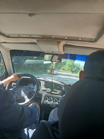 An interior shot from inside a car shows a driver's hands on the steering wheel as the vehicle travels on a road. The driver is wearing a dark suit jacket and is seated in the driver's seat of what appears to be a Volkswagen. The dashboard is visible, including the steering wheel with the Volkswagen logo, an instrument cluster with a speedometer, and a radio. A passenger is seated in the foreground, with their head and shoulders visible from the back, wearing a black shirt. The road outside is lined with trees, and another car is visible in the distance. The scene is lit by daylight. The image is believed to be taken in Talgar, Kazakhstan.