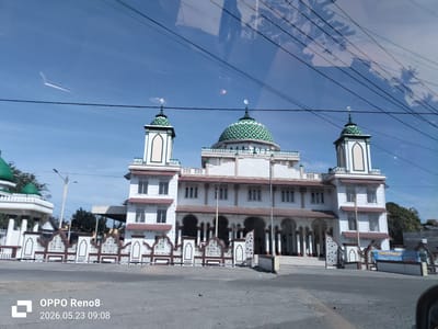 The image shows a mosque located in Dua Paya, Indonesia. The mosque is a two-story building painted white with a green and white patterned dome and two smaller towers on either side. The dome and towers are topped with crescent moon symbols. The building has arched entrances supported by white pillars. In front of the mosque is a decorative fence made of white and brown panels. The sky is blue with a few power lines visible across the top of the frame. There are reflections in the windshield of the car taking the photo. A small white structure with a green dome can be seen to the left. A banner or sign is on the right. The date and time on the lower left corner indicate the photo was taken with an OPPO Reno8 on May 23, 2026, at 09:08.