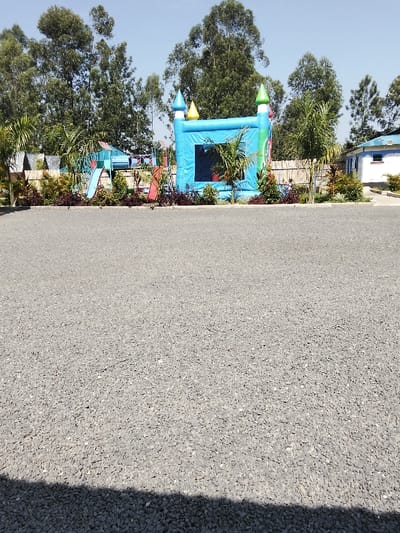 The image displays an outdoor recreational area located in Malaha Isongo Makunga ward, Kenya, viewed under a clear blue sky. The foreground is a large expanse of light grey gravel or crushed stone, with a dark shadow occupying the bottom margin. A low, manicured garden bed containing dark red and green shrubs forms a boundary beyond the gravel. Behind this border, a children's play area is visible. This area features a prominent, inflated blue bounce house shaped like a castle with turrets colored blue, yellow, and green. To the left of the bounce house, a blue playground structure with a slide is partially visible. Several slender palm trees are positioned throughout the play area. A light-colored wooden fence demarcates the rear of the playground. In the far right background, a white building with a blue roof and blue window frames is present. The background is dominated by a dense line of tall, leafy green trees that extend upwards to the horizon. No individuals are present in the scene.