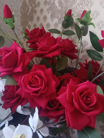 A close-up, eye-level shot captures a bouquet of artificial red roses and white flowers arranged in a vase against a beige patterned wallpaper. The red roses, with their velvety petals and green leaves, are the dominant element, with some buds still closed and others in full bloom. White orchid-like flowers with yellow centers peek out from the bottom of the arrangement. The wallpaper behind the bouquet features an ornate, damask-like pattern in shades of beige and gold, creating a decorative backdrop. There are no people present in the image. The scene appears to be an indoor setting, possibly a home or a commercial establishment. The lighting is soft and even, highlighting the textures of the flowers and wallpaper. The image does not contain any geographical markers or specific details that would allow for its localization in Tallinn, Armenia or any other city.
