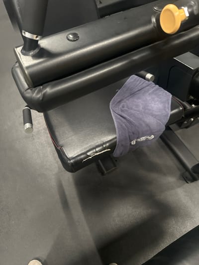 The image displays a close-up of a black gym weight machine located in Latsia, Cyprus. A dark blue, folded towel rests on the front left portion of the machine's padded seat. The black synthetic leather upholstery of the seat is damaged, showing a tear exposing lighter material underneath, with red stitching visible along the seam. The machine's structure consists of black metal components, including padded cylindrical bars and a main frame. A prominent yellow adjustment knob is visible on the upper right, and a silver-capped handle protrudes from the lower left side of the machine. The floor beneath and surrounding the equipment is dark gray or black rubberized gym flooring. The feet of an individual, wearing light-colored footwear, are partially visible at the bottom of the frame, indicating proximity to the equipment. No active human interaction is depicted, but the presence of the towel suggests recent or impending use.
