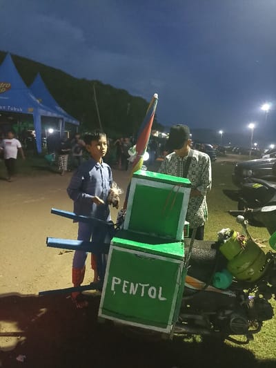 This image captures an outdoor evening scene, likely a market or event, in Kecamatan Pujut, Indonesia. In the foreground, a young boy stands on the left, wearing a blue shirt and trousers with distinctive red footwear, holding a small package or bag of food.
To his right is a green street food vendor cart with "PENTOL" written in white letters on its side. An open colorful umbrella (red, blue, yellow stripes) is attached to the cart. The cart is mounted on a motorcycle, and a small green gas cylinder is visible on the back of the motorcycle, presumably for cooking or heating the food. Partially obscured by the cart and the umbrella, an adult male wearing a patterned shirt and a dark cap is attending to the vendor stall.
In the background, several other people are visible, some gathered near blue tents. One tent displays text, including "Hydro Coco" and "iun Tubuh." The sky is dark, indicating evening or night, with distant hills or mountains silhouetted against it. Bright streetlights illuminate the area in the mid-ground and background, and several vehicles are parked further away. The ground consists of dirt and patches of grass. The scene portrays a casual, communal atmosphere at an outdoor gathering or market.