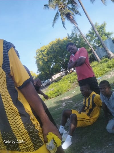 The image shows a group of young men standing and sitting on a grassy area in Nungwi, Tanzania, located at latitude -5.726 and longitude 39.301. One man is standing with arms crossed wearing a red polo shirt and black shorts. Another man is standing wearing a black and yellow shirt with black pants, and he is holding a yellow cloth in his hand. There is a third man sitting on the ground, also wearing a black and yellow shirt and yellow shorts. He is looking down at the ground. A fourth man is seated behind the third man, looking directly at the camera. The fourth man is wearing a grey shirt and black shorts. A palm tree and another smaller tree are visible in the background. There are tall buildings in the distance, to the right of the frame. The sky is blue with a few white clouds. The image was taken during the day and the lighting is bright.