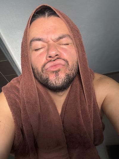 An angled, close-up shot depicts a person, likely male, wrapped in a brown, textured towel that covers their head and upper body. The person's face is central to the image, showing a strong, contorted expression. Both eyes are squeezed tightly shut, and the lips are pursed and pushed forward, conveying a sense of discomfort, pain, or intense sensory experience, possibly in an exaggerated or playful manner. Dark hair is visible around the forehead and sideburns, along with a short beard or stubble on the cheeks and chin.
Water droplets are clearly visible on the person's skin, particularly on the shoulder and upper arm, suggesting they have recently showered or are undergoing a water-based treatment. The background consists of a plain, light gray wall on the left side and a darker, possibly tiled, surface on the right, indicating an indoor setting like a bathroom or spa. The lighting is uniform and soft. No text is visible in the image. The scene captures a moment of heightened physical sensation, set in San Juan Cuautlancingo, Mexico.